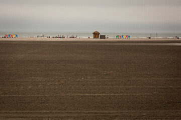 Long view of the brown sand on a beach during low tide with people, cabanas, and lockers along the ocean shore in the distance on an overcast day. 