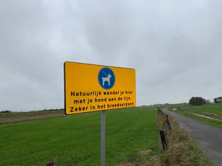 Fototapeten Straßenschild in niederländischer Sprache. Gehen Sie hier mit Ihrem Hund an der Leine spazieren, besonders in der Brutzeit. (Natürlich gehen Sie hier mit Ihrem Hund an der Leine, besonders in der Brutzeit). Marken / Niederlande. © Lukas
