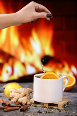 A woman's hand puts a slice of orange into white tea cup with lemon, ginger, orange and jam against the background of a burning fireplace. Healthcare. Vitamin cocktail.