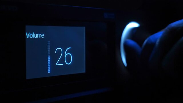 In the dark, the hand turns the volume control, illuminated by blue light on the front panel of the black stereo equipment. The display shows the music volume level. Closeup