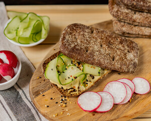 Healthy vegan sandwich - burger close up with cucumber, hummus and radish on whole grain bread, wooden cutting board. Cooking ingredients, diet concept. Selective focus, copy space. Organic products.