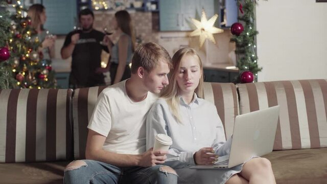 Young Freelancer Couple Working Online While Group Friends Have Fun Celebrate Christmas. Girl And Man Uses Computer. Students Preparing For Exams At University In Dormitory. Xmas New Year Holiday.