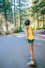 Teen girl in helmet skateboarding on road through forest 