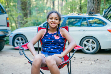 Obraz premium Happy smiling girl sitting in camping chair in campground in forest