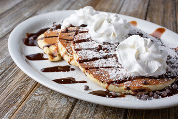 A view of a plate of chocolate chip pancakes, featuring whipped cream and powdered sugar toppings, in a restaurant or kitchen setting.