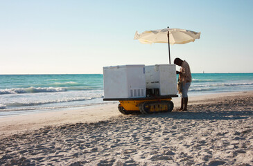anonymous summer ice cream man with his cart on the sandy beach rests under the umbrella of his...