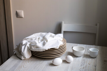 a stack of plates with an embroidered crumpled napkin, two eggs and two empty tea cups on a gray wooden kitchen table