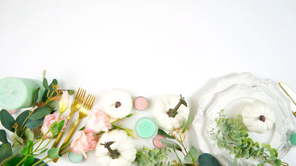 Happy Thanksgiving table setting with white vintage style plates, gold silverware, and centerpiece with greenery, pink, and white pumpkins, on white tablecloth. Top down view flat lay with copy space.