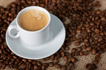 White cup of frothy coffee and sprinkled beans on sacking. Close-up. Top view.