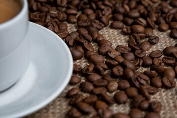 Scattered roasted coffee beans and a white cup with a drink on sacking. Close-up. Top view.
