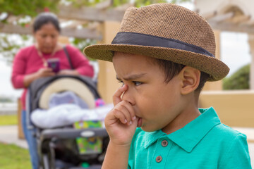 An upset toddler wearing a Fedora hat and thumb in his mouth while a lady behind him is texting....