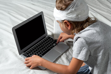 Little Caucasian blond boy with a head injury and bandage is sitting on the bed and using laptop. Recovery after incident. Remote communication, distance learning, technologies and gadgets in life