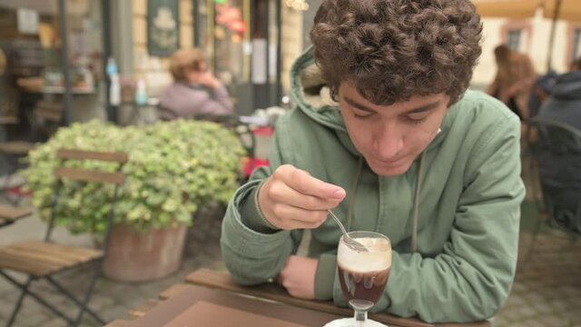 A Caucasian boy consumes his chocolate at an outdoor table. He has chosen a place far from the others to avoid contact, has the mask to defend himself from the coronavirus is lowered under his chin.