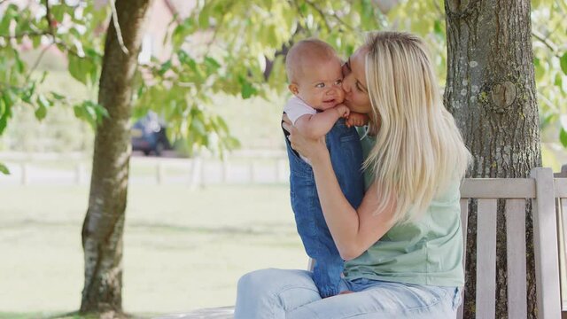 Loving mother with baby daughter sitting on seat under tree in summer park together - shot in slow motion