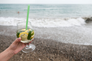 White female hand holding a wine glass with tropical mojito cocktail. Recreational getaway or romantic holiday on remote island. Luxury drink on a beach near the sea. Waves and sand in background.