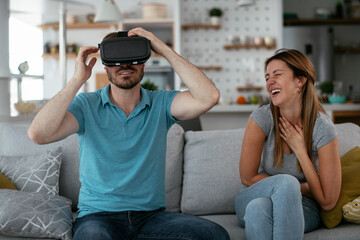 Smiling young man using VR headset at home on couch. Woman and her husband enjoying virtual reality in apartment..