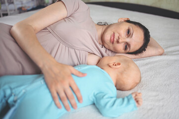 Obraz na płótnie Canvas Young mother lies with newborn infant baby in blue jumpsuit on bed and breastfeeds him breast milk