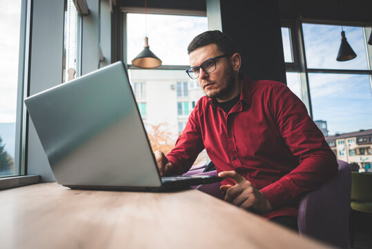Freelancer man with face mask in red shirt with glasses working on laptop in coffee shop, remote work