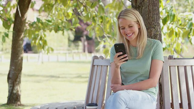 Woman sitting and relaxing on park bench under tree in summer using mobile phone to browse internet and social media - shot in slow motion