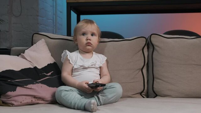 Cute baby girl is sitting on gray sofa in white T-shirt and blue pants with black remote control in her hands and watching TV. Kid watches cartoons on TV.
