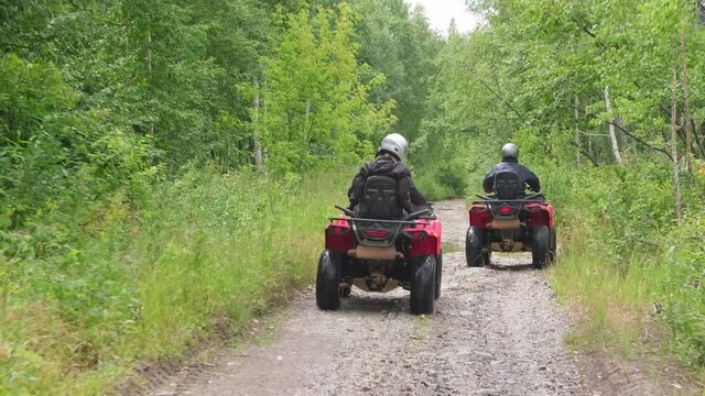 Tracking shot of people in riding gear driving red quad bikes along narrow forest road in rough terrain