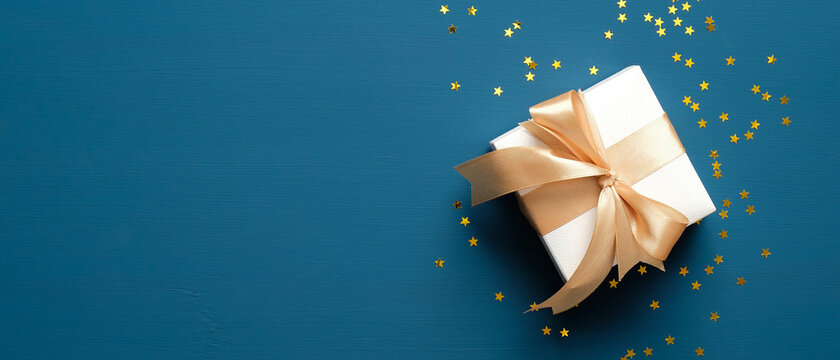 White gift box with golden ribbon bow on blue background with confetti. Christmas present, valentine day surprise, birthday concept. Flat lay, top view.