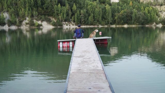 4K Man with blue backpack wearing cap with visor backwards sitting on pier with his beagle. Lifestyle concept, animals concept.