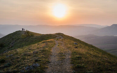 The road along the mountain ridge with silhouettes of people on the background of sunset against the mountains in the fog
