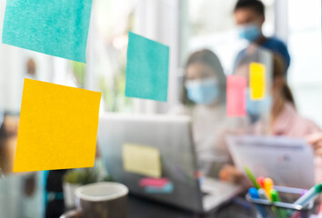 Post-it paper or remind paper for writing text on clear acrylic partitions In the Home Office with blur group of colleagues are consulting or brainstrom, Soft focus and focus at foreground.