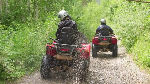 Tracking shot of people in helmets riding quad bikes through puddles along narrow forest road in rough terrain
