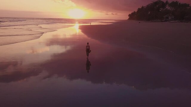 Rear extreme long tracking of unrecognizable female person walking along extensive coast. Landscape of endless ocean view from tropical island, sun setting over horizon, sky reflecting on wet sand