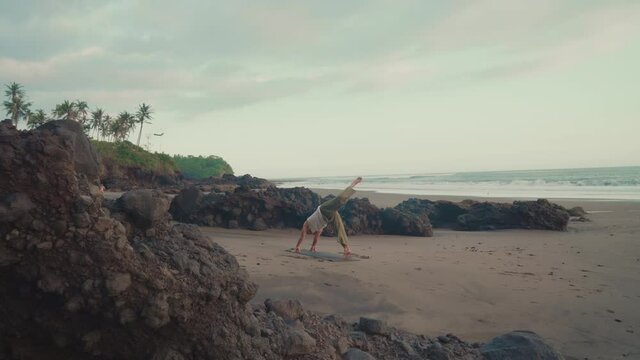 Wide rear shot of middle-aged fit male ascetic practicing yoga on mat placed on sandy ocean coast among rocks. Man doing sports outdoors on island of Bali