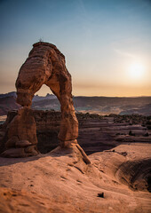 View of the delicate arch in arches national park at sunset. The sun can be seen and the land around it is lit up from the sunset. 