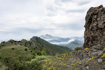 The slope of the mountain with yellow flowers and a vertical stone column against the mountains in the clouds