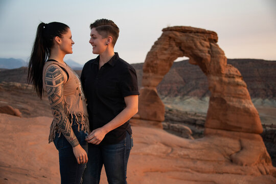 Lesbian couple smiling at each other in front of the Delicate Arch in Arches National Park. The sun is setting which is giving the couple a nice yellowish glow.