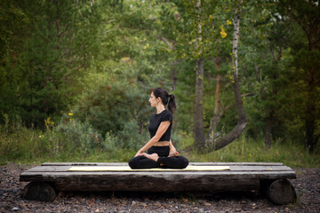 a woman in a black suit does yoga in the woods on a wooden pedestal
