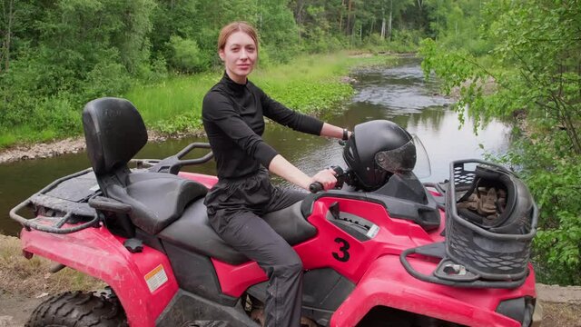 Portrait of cheerful woman sitting on red quad bike in forest by the river and smiling for camera