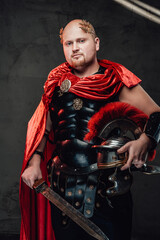 Fototapeta na wymiar Royal roman soldier with bald head dressed in armour and red cape poses holding a sword and helmet in dark background.