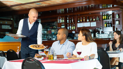 Smiling waiter serving delicious pizza to couple in cozy restaurant