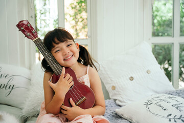 Asian little girl with wooden classic ukulele on sofa at home.