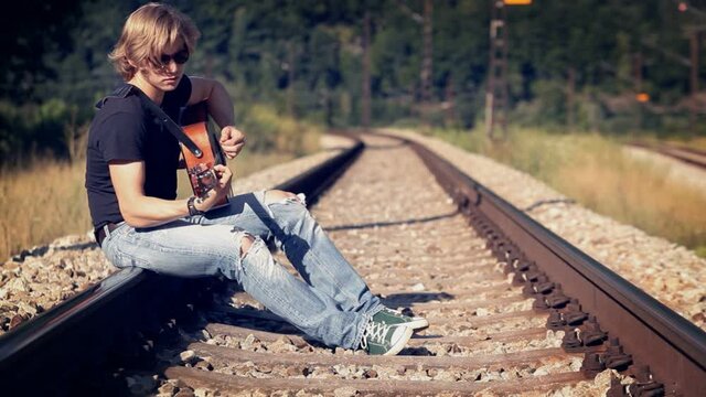 Long-haired teenage guitarist sitting on railway tracks strumming strings on acoustic guitar. Live music performance, hobo style, adolescence concept.