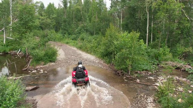 Aerial tracking of people in helmets driving red ATV through mud and puddles on forest road