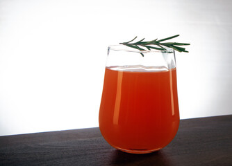Semicircular glass of fresh orange juice with rosemary sprig on top staying on a wooden table against light background