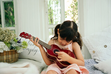 Asian little girl with wooden classic ukulele on sofa at home.