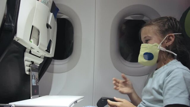 Little girl caucasian at plane with wearing protective medical mask. Child baby tourist at aircraft with respirator play at smartphone mobile phone. Coronavirus epidemic sars-cov-2 covid-19 2019-ncov.
