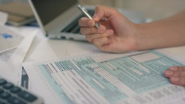 Close up of unrecognizable female hand writing in printed tax return form completing information with blurred laptop and papers in background