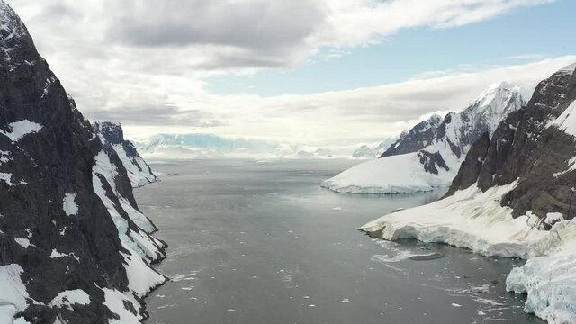 4K Aerial Landscape of snowy mountains and icy shores in Antarctica. Beautiful blue iceberg with mirror reflection floats in open ocean. Glaciers of a harsh continent.