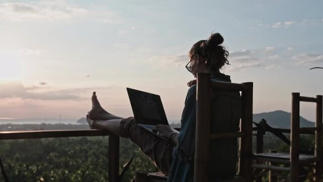 Woman freelancer sitting with a laptop on a high bar chair. Amazing sunset-view and thick jungle near the blue sea.