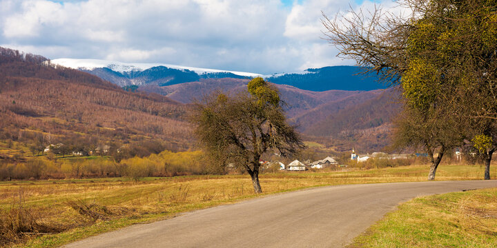 old road through countryside in early spring. leafless trees along the way. snow capped mountain in the distance. sunny weather with clouds on the sky