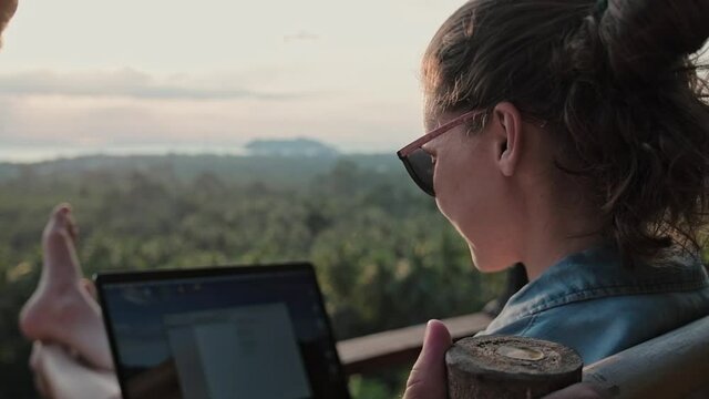 Freelancer woman sits with a laptop on a high bar stool. Amazing sunset views and dense jungle by the blue sea.